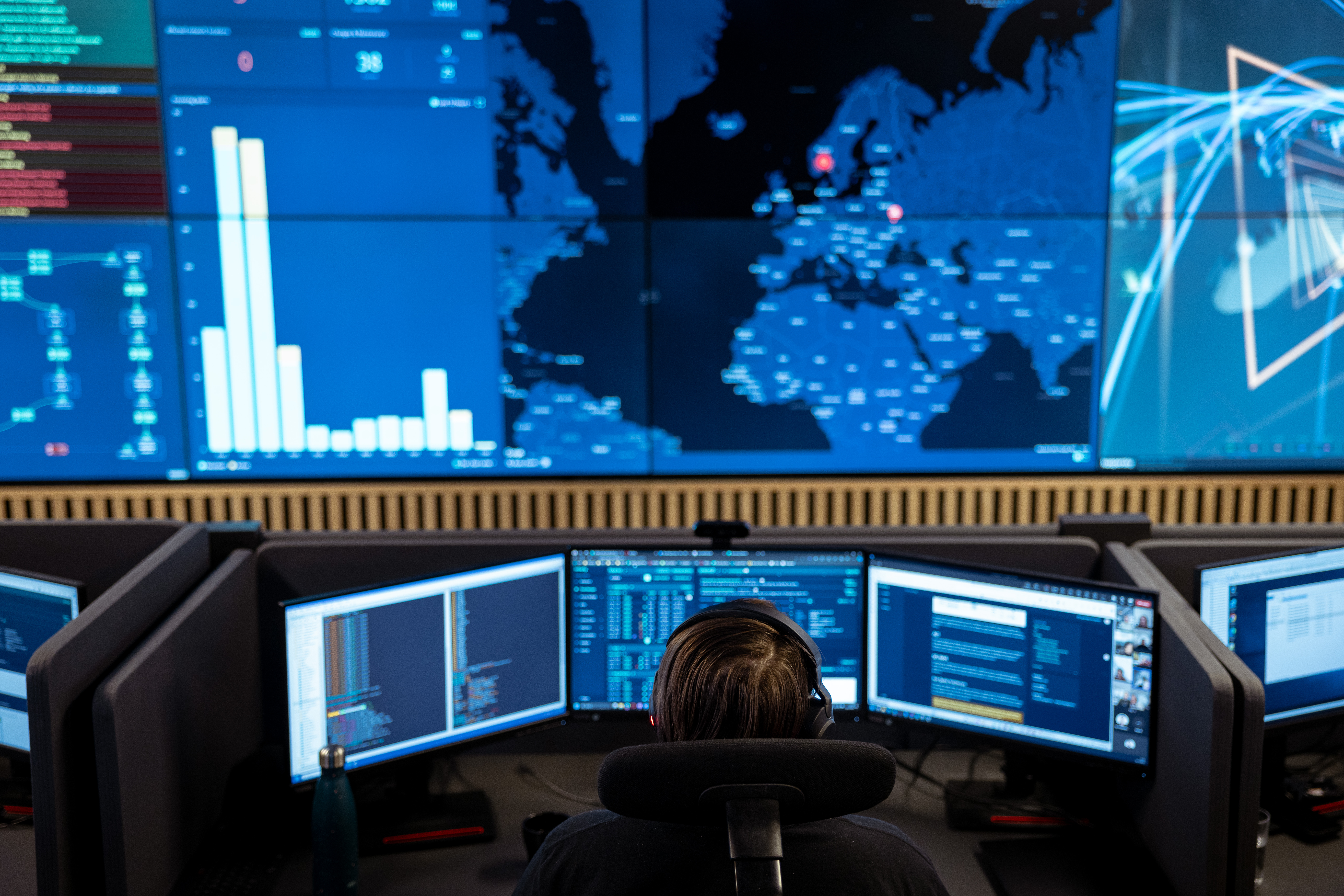 Person working at multiple computer monitors in a control room with a large digital world map and data charts on the wall.